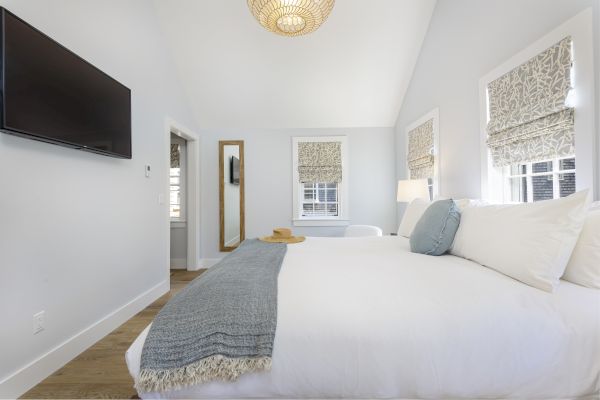 A serene bedroom with a neatly made bed, wall-mounted TV, full-length mirror, and windows with patterned shades in a bright, airy space.