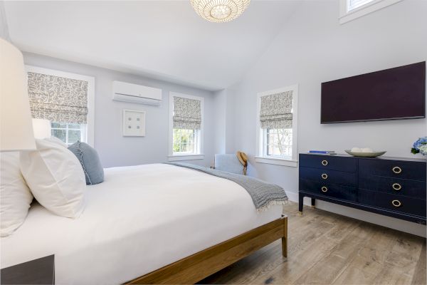 A modern bedroom with a bed, pillows, and a blanket. There's a dresser with a TV above, wall art, windows with blinds, and an air conditioner.