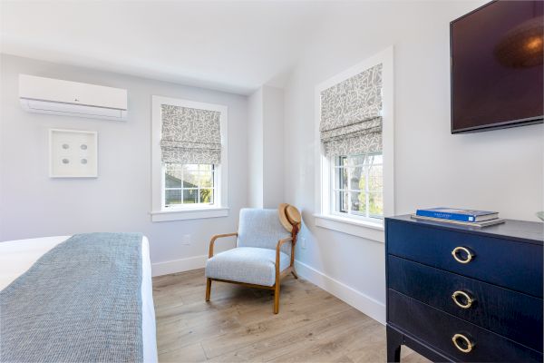 A bright bedroom with a chair, dresser, window treatments, wall art, and an air conditioning unit. It's neatly organized and well-lit, creating a cozy vibe.