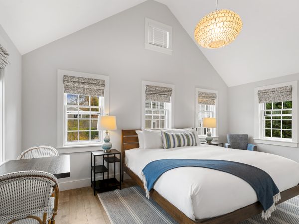 A cozy bedroom with a large bed, blue throw, chairs, lamps, a desk, and several windows with blinds.
