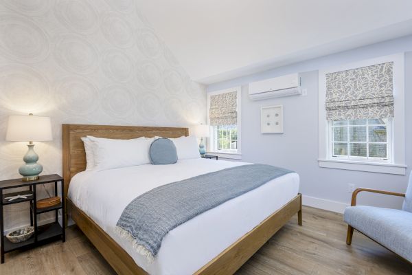 A modern bedroom with a wooden bed, gray blanket, pillows, side table with lamp, a/c unit, armchair, and patterned window shades.