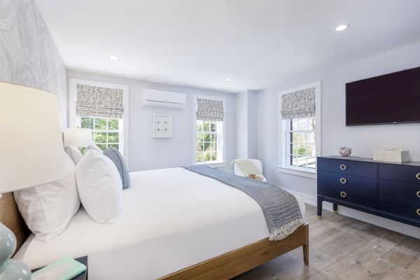 A neatly arranged bedroom with a double bed, three windows, a wall-mounted TV, a dresser, and a small chair, all in a bright and airy setting.