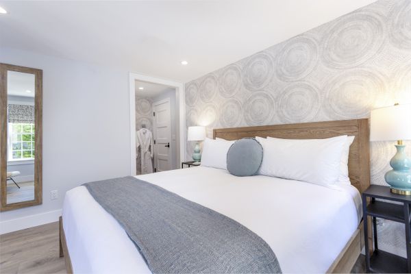 A neatly arranged bedroom featuring a bed with white sheets and a gray blanket, a bedside lamp, a large mirror, and a doorway leading to a bathroom.