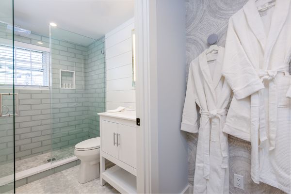 A modern bathroom with a glass shower, gray subway tiles, a white vanity, a toilet, and two white robes hanging on hooks on the wall.