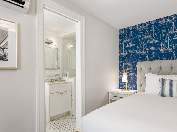 A modern bedroom with a blue nautical-themed accent wall, framed boat picture, and attached bathroom featuring a white vanity and shower.