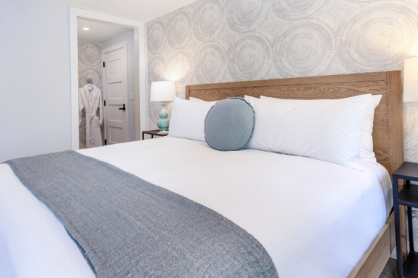 A neatly made bed with white linens, a light blue runner, a round pillow, bedside tables, lamps, and modern wallpaper. A door leads to another room.