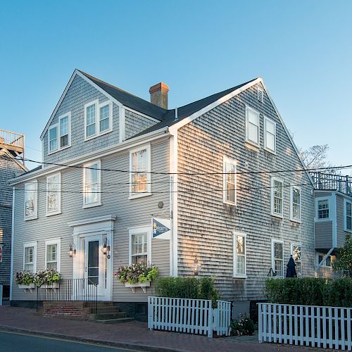 A grey wooden house with a white picket fence, flower boxes, and neighboring buildings bathed in daylight on a quiet street.