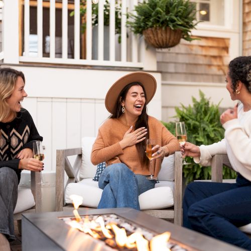 Three people are sitting outdoors around a fire pit, laughing and holding drinks, with plants and a house in the background.