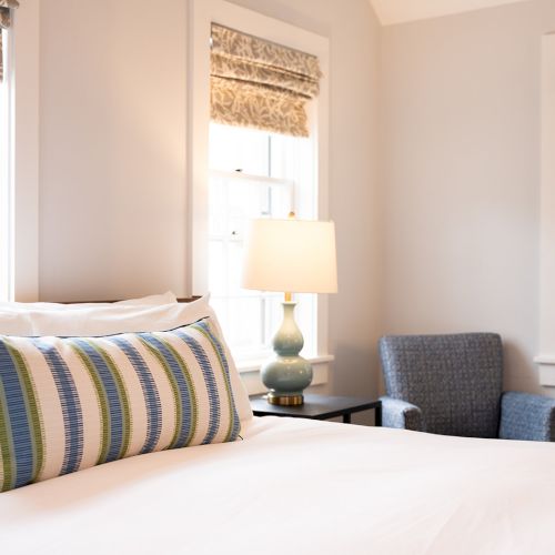 A cozy bedroom with a bed, striped pillow, lamp, and an armchair near windows. Light filters through the patterned window shades.