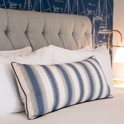 A neatly made bed with a striped pillow, tufted headboard, nautical wallpaper, and a lit bedside lamp is shown.