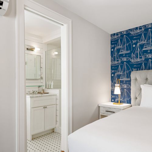 A cozy bedroom with a blue nautical-themed wall, white bedding, and a view of a small bathroom with a white vanity and patterned floor.