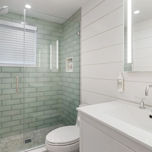 A modern bathroom with a glass shower, light green tiles, a white sink cabinet, and a wall-mounted mirror.