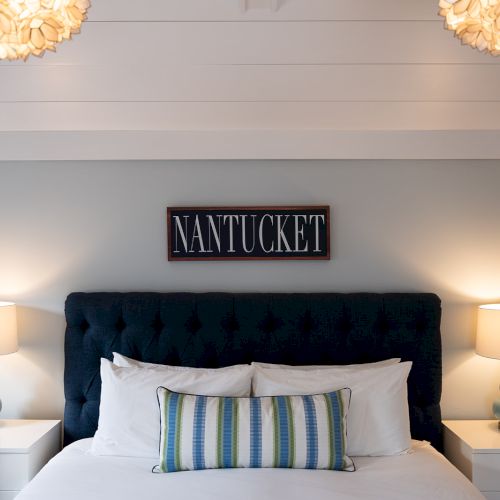 A cozy bedroom with a "Nantucket" sign, tufted headboard, striped pillow, two nightstands, and lamps under floral ceiling lights.