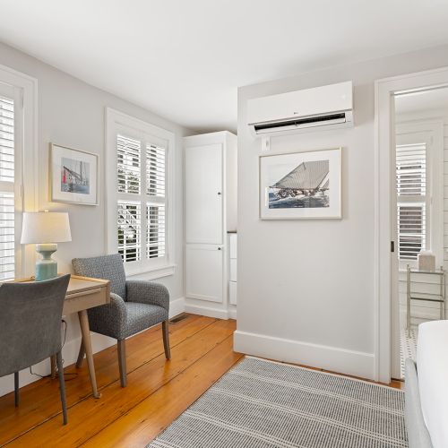 A bright room features a desk, chairs, wooden floor, and an adjacent bathroom, with white walls and large windows completing the scene.