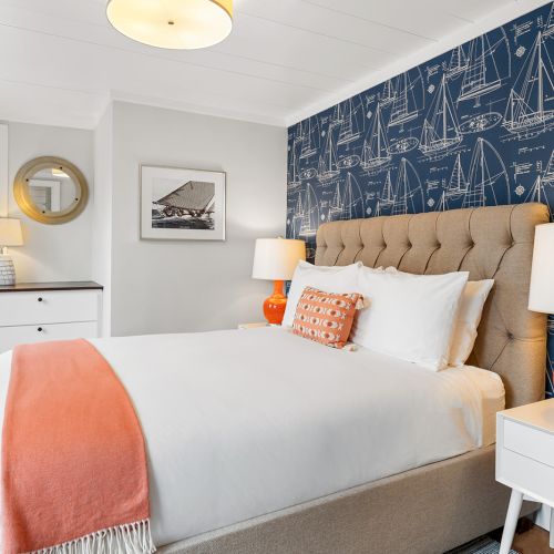 A cozy bedroom features a bed with white and orange accents, nautical-themed wallpaper, and stylish lamps, creating a warm ambiance.