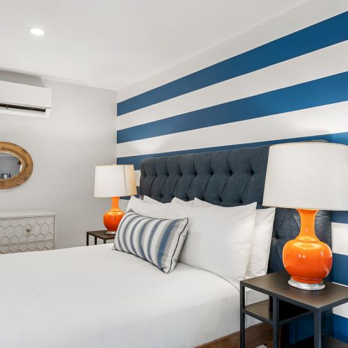 A bedroom with a striped blue and white wall, a bed with white linens, orange lamps, and a mirror above a dresser.