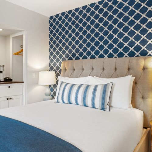 A cozy bedroom with a patterned blue accent wall, tufted headboard, and blue accents. There's a side table with a lamp and a closet.