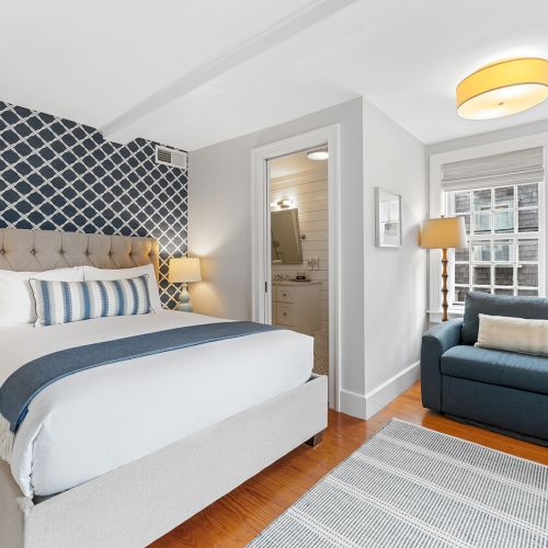 A cozy bedroom with a bed, patterned wall, sofa, rug, two lamps, and a wall-mounted TV, next to a bathroom.