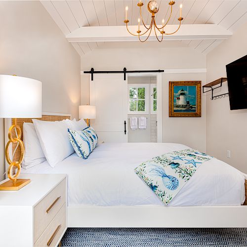 A bright bedroom features a bed with white linens, blue accent pillows, a wooden nightstand, a chandelier, a flat-screen TV, and a sliding barn door.