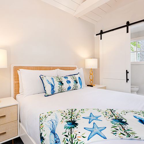 A cozy bedroom with a bed adorned with aquatic-themed bedding, two nightstands with golden lamps, and a sliding barn door leading to the bathroom.