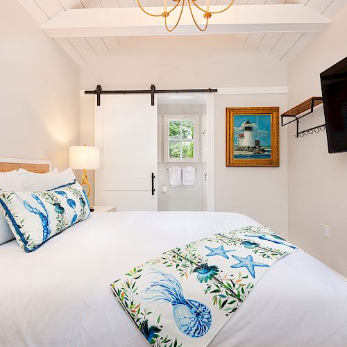 A cozy bedroom with white walls, a bed with blue and sea-themed decor, a TV on the wall, a lamp, and a framed picture of a lighthouse.