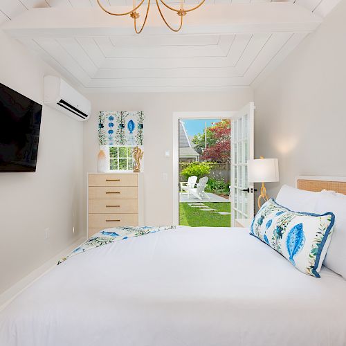 A cozy bedroom with a large TV, dresser, air conditioner, and white bed. French doors open to a garden with chairs and a lamp stands nearby.