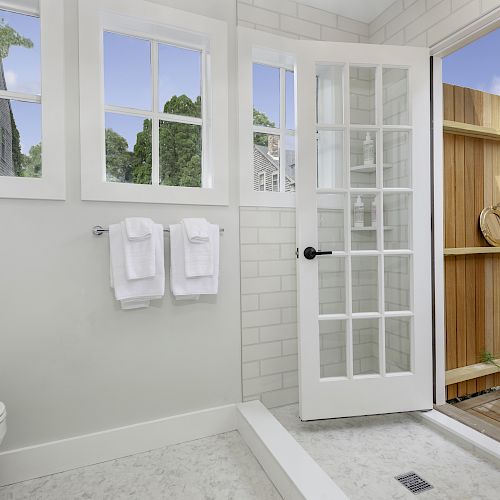 A bright bathroom with a toilet, two towels on the wall, and a door leading to an outdoor area with wooden walls and potted plants.