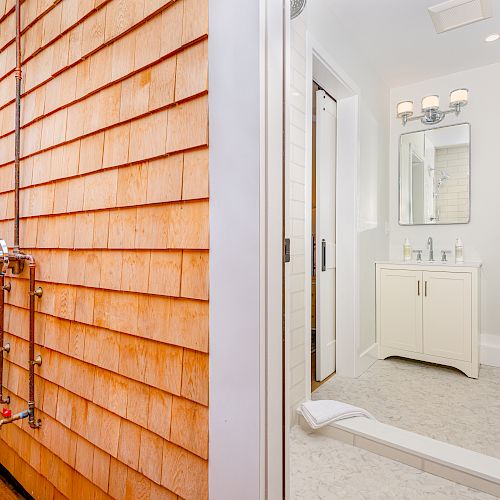 The image depicts a bathroom with a toilet, sink, and mirror on the right side. An outdoor shower area with wooden siding is visible on the left side.