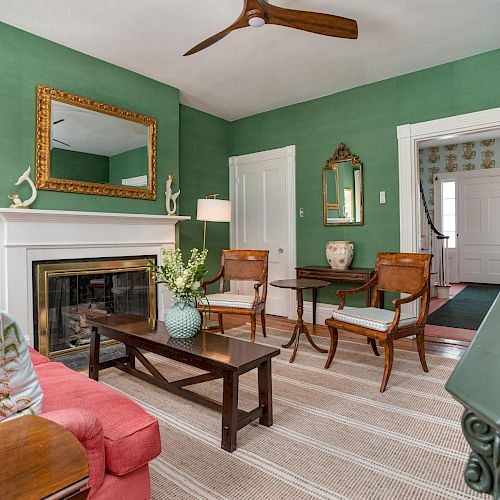 A living room with green walls, a red sofa, a fireplace, wooden chairs, a ceiling fan, and various decor items, including a mirror and paintings.