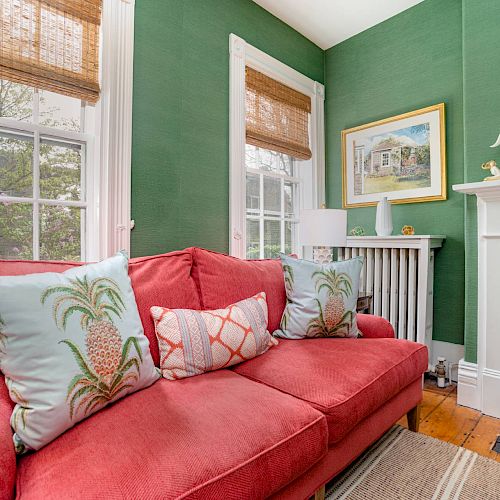 A cozy living room with green walls, a red sofa adorned with patterned pillows, white-framed windows, and a white fireplace decorated with artwork.