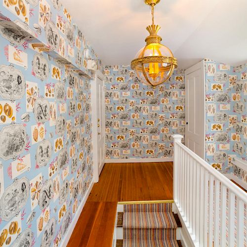 A small hallway with blue, patterned wallpaper featuring various illustrations, a wooden floor, a white railing, and a golden chandelier.