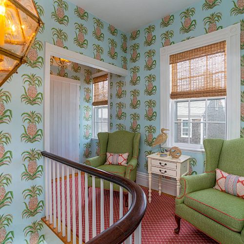 The image shows a room with pineapple-patterned wallpaper, green armchairs, a white side table, bamboo blinds on windows, and a hanging lantern light.