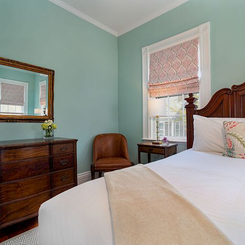 This image shows a cozy bedroom with a wooden dresser, a framed mirror, a chair, a bedside table, and a bed with pillows and a throw.