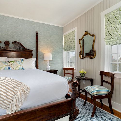 A cozy bedroom features a large wooden bed, two chairs, a side table with a lamp, decorative mirrors, and light-colored window blinds.