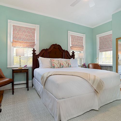 A cozy bedroom with a large bed, wooden headboard, green walls, dresser, mirror, and two windows with shades.