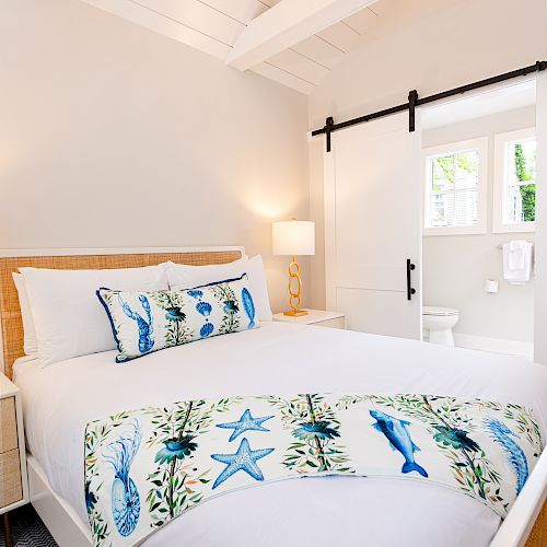 A cozy bedroom with a white bed featuring ocean-themed pillows and throw, a bedside lamp, and a sliding barn door leading to a bright en-suite bathroom.