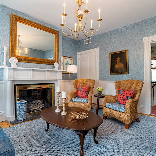 A cozy living room with a blue theme featuring wicker chairs, a fireplace, ornate mirror, chandeliers, and wall art. Door opens to another room.