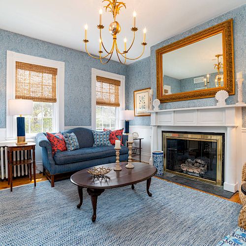 A cozy living room with blue walls, a chandelier, a large mirror above a fireplace, two windows, a sofa, chairs, and a coffee table ending the sentence.