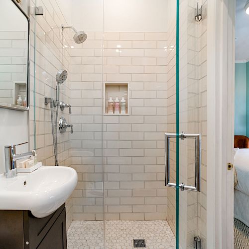 A bathroom with a glass shower, white tile walls, a wall-mounted sink, and a glimpse of a bedroom with a bed and window in the background.