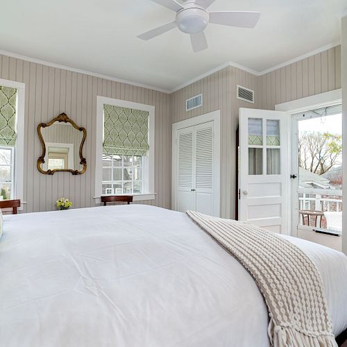 This image shows a cozy bedroom with a large bed, white linens, decorative pillows, patterned blinds, a ceiling fan, and a wall-mounted TV.