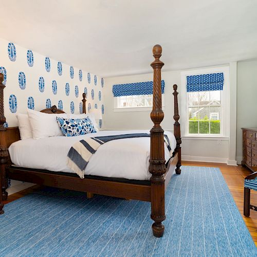 A bedroom with a four-poster bed, blue accents, a patterned wall, wooden furniture, a TV, and natural light coming from the windows.