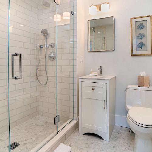 The image shows a modern bathroom with a glass-enclosed shower, toilet, single-sink vanity, and wall-mounted mirror, all in a light color scheme.