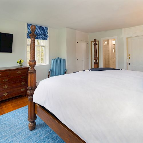 The image shows a bedroom with a large bed, wooden dresser, wall-mounted TV, blue rug, and window with blue curtains, leading to a bathroom.