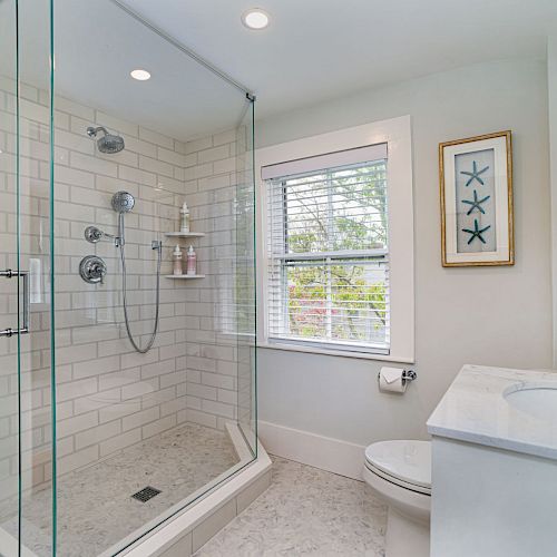 A modern bathroom with a glass-enclosed shower, white tile, window with blinds, toilet, sink with countertop, and framed starfish art.