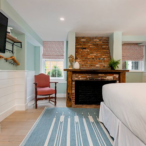 A cozy bedroom with a fireplace, TV, red chair, and bed with white bedding. Decor includes plants and curtains; light blue rug on the floor.