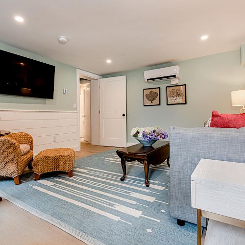 A cozy living room with a TV, wicker chairs, a blue-striped rug, a gray sofa, wall decor, and bright lighting.