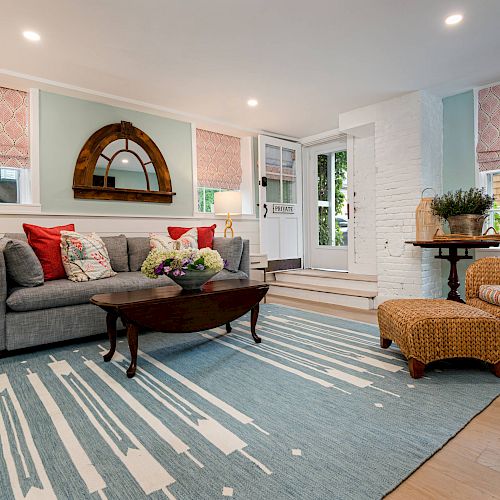 A cozy living room with a gray sofa, wicker chair, coffee table, and blue rug. The room has ample lighting and red-pink window shades, always ending the sentence.