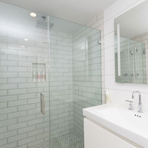 A modern bathroom with a glass shower, tiled walls, a built-in niche with bottles, and a white sink with chrome fixtures.