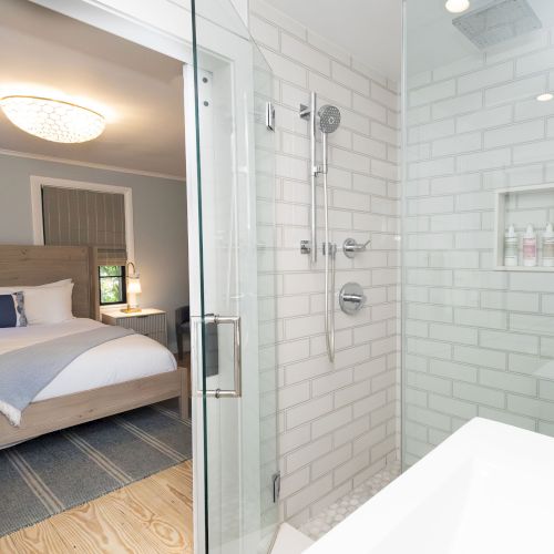 A modern en-suite bathroom with a glass shower, chrome fixtures, and white subway tiles beside a cozy bedroom with a bed and lamp.