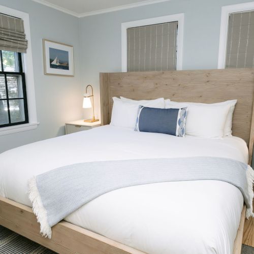 A bright bedroom with a wooden bed, white linens, blue pillows, two window blinds, and bedside lamps.